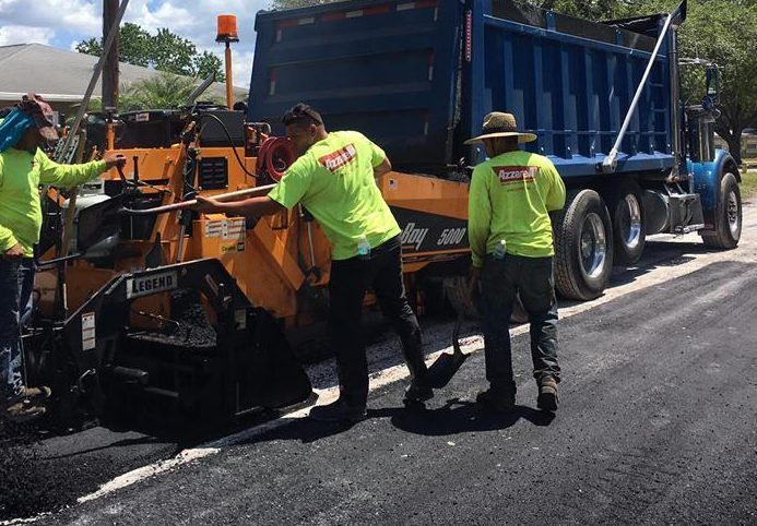 Road construction workers in lime green shirts paving a road with a truck in a residential area.