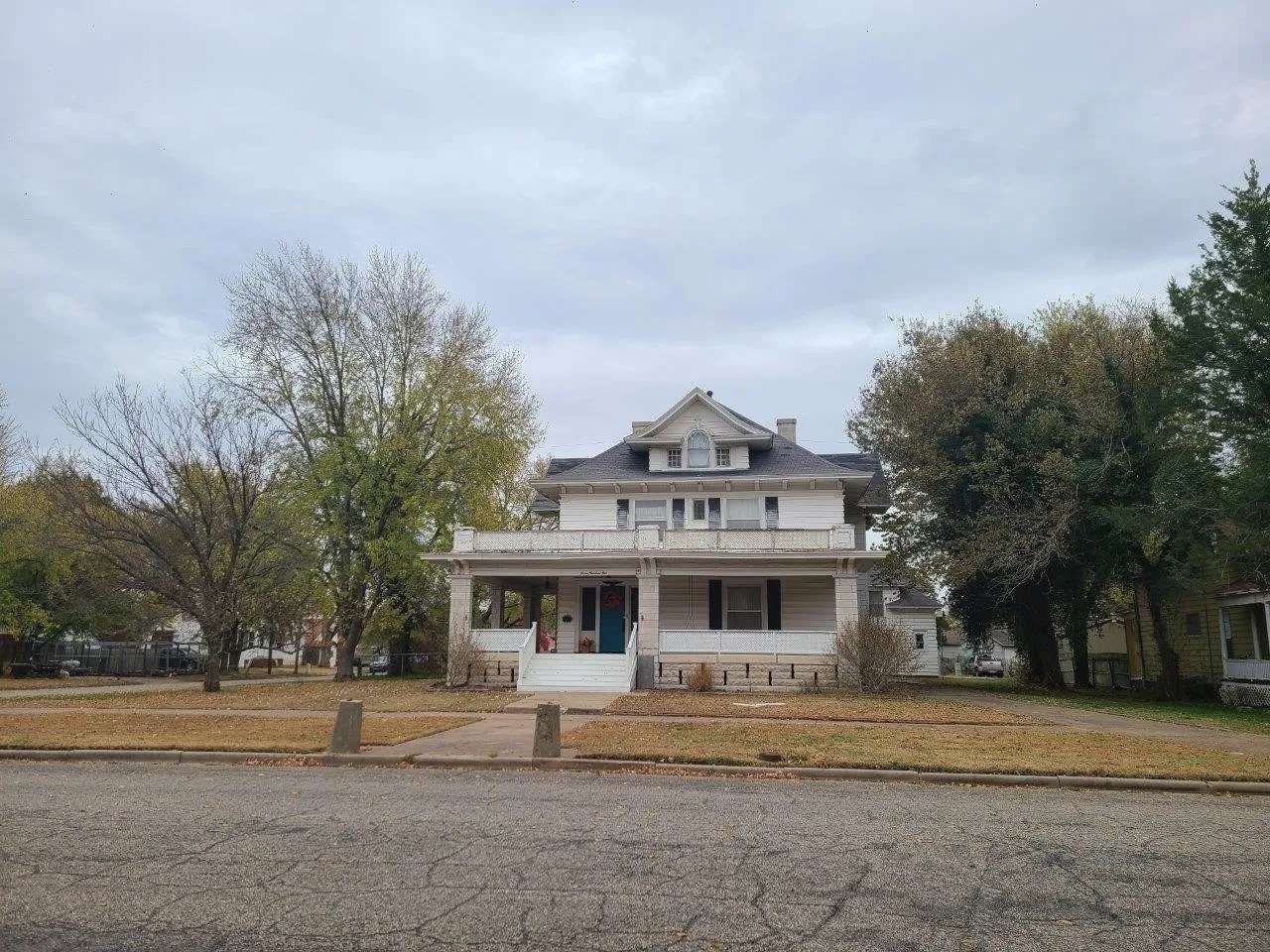 Old white farmhouse with a front porch and trees, viewed from a gravel road under a cloudy sky