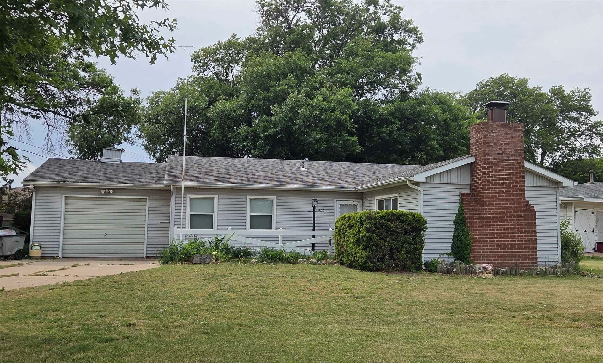 Single-story white house with a red brick chimney and overgrown front lawn under cloudy skies