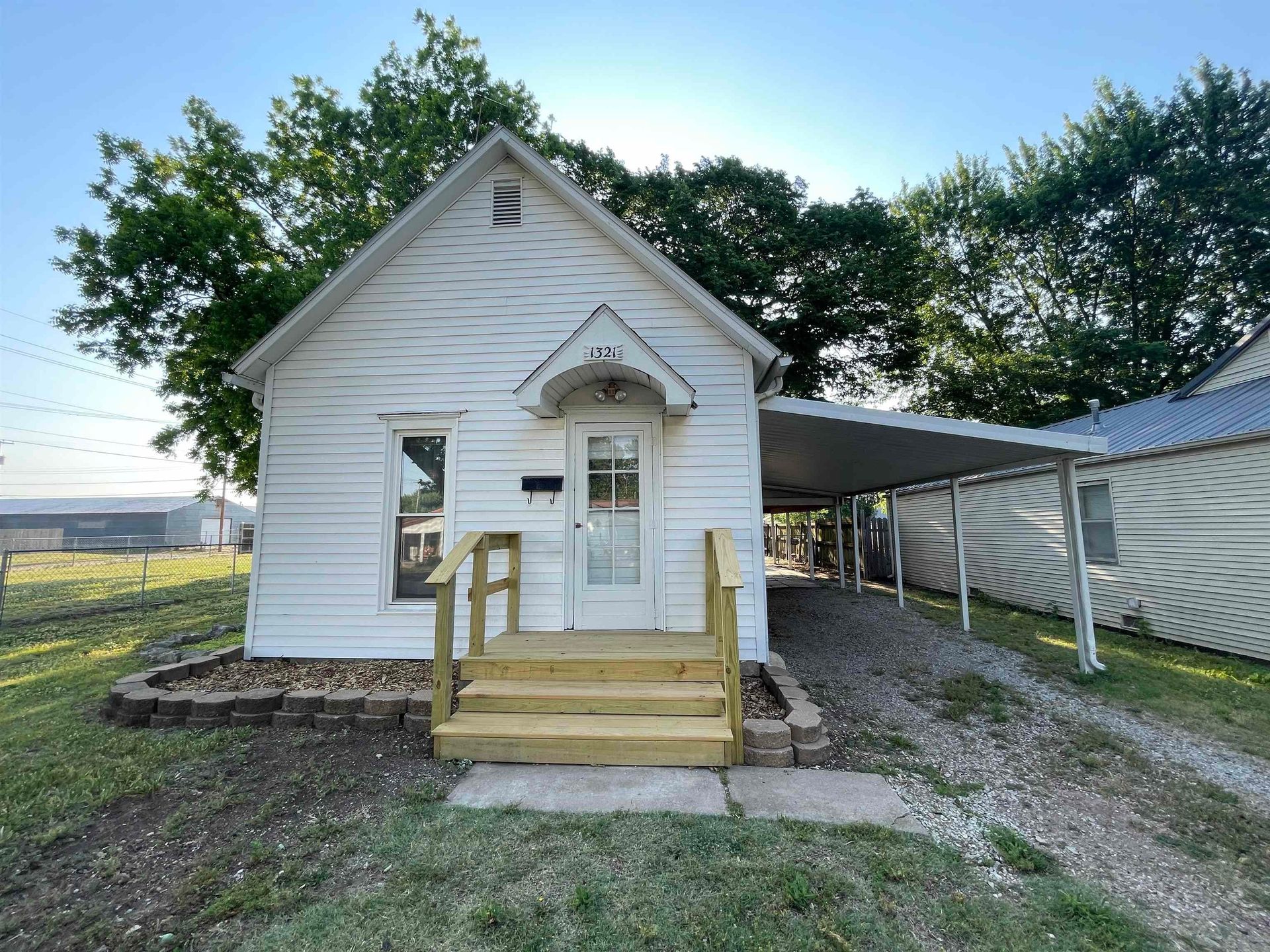 Small white house with front steps, gravel driveway, and a carport beside it under a clear sky