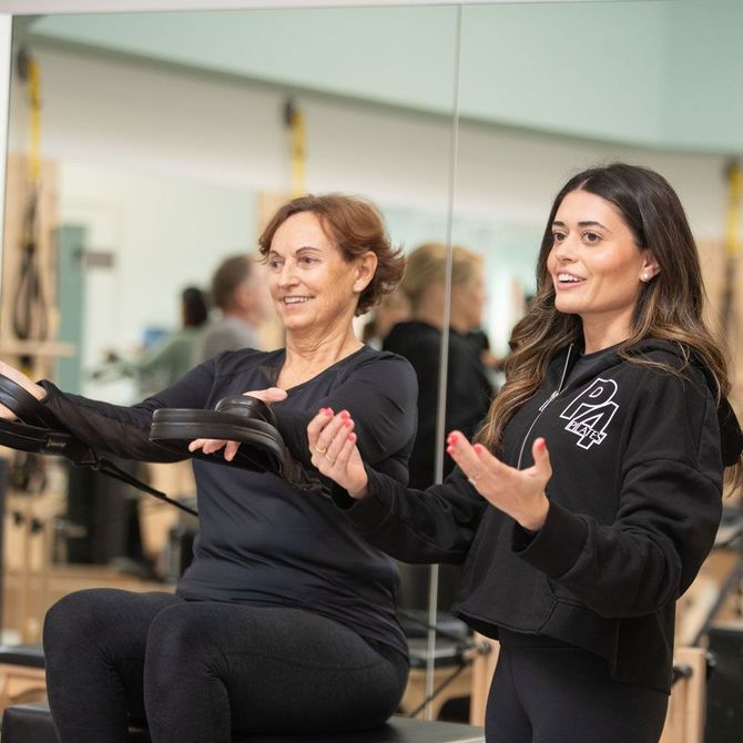 Two women are sitting on a pilates machine in front of a mirror.