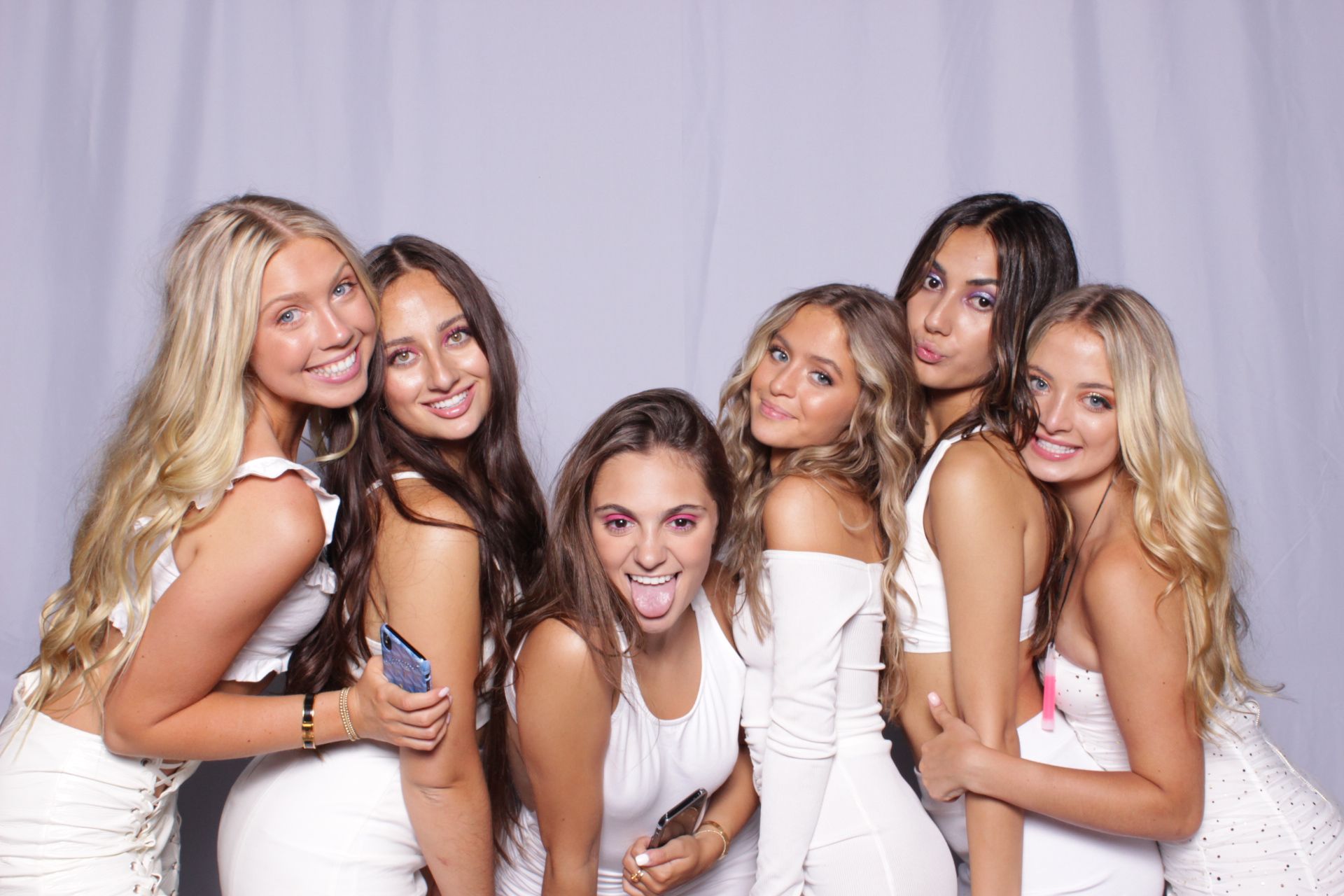 Group of seven women in white outfits smiling in a photo booth, posing against a light gray backdrop.