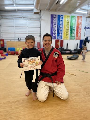 A man in a red karate uniform is kneeling next to a young boy holding a certificate.