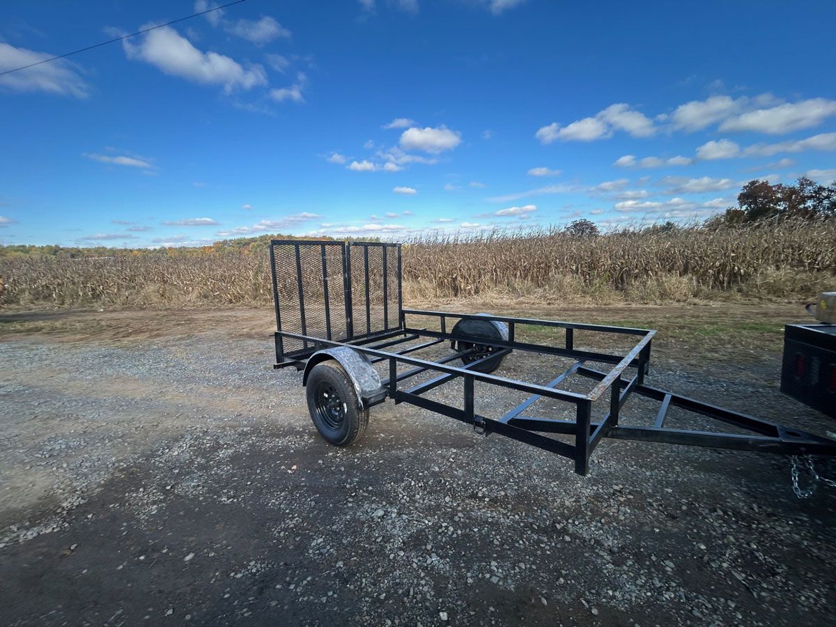 Black utility trailer on gravel with a dry field and blue sky in the background.