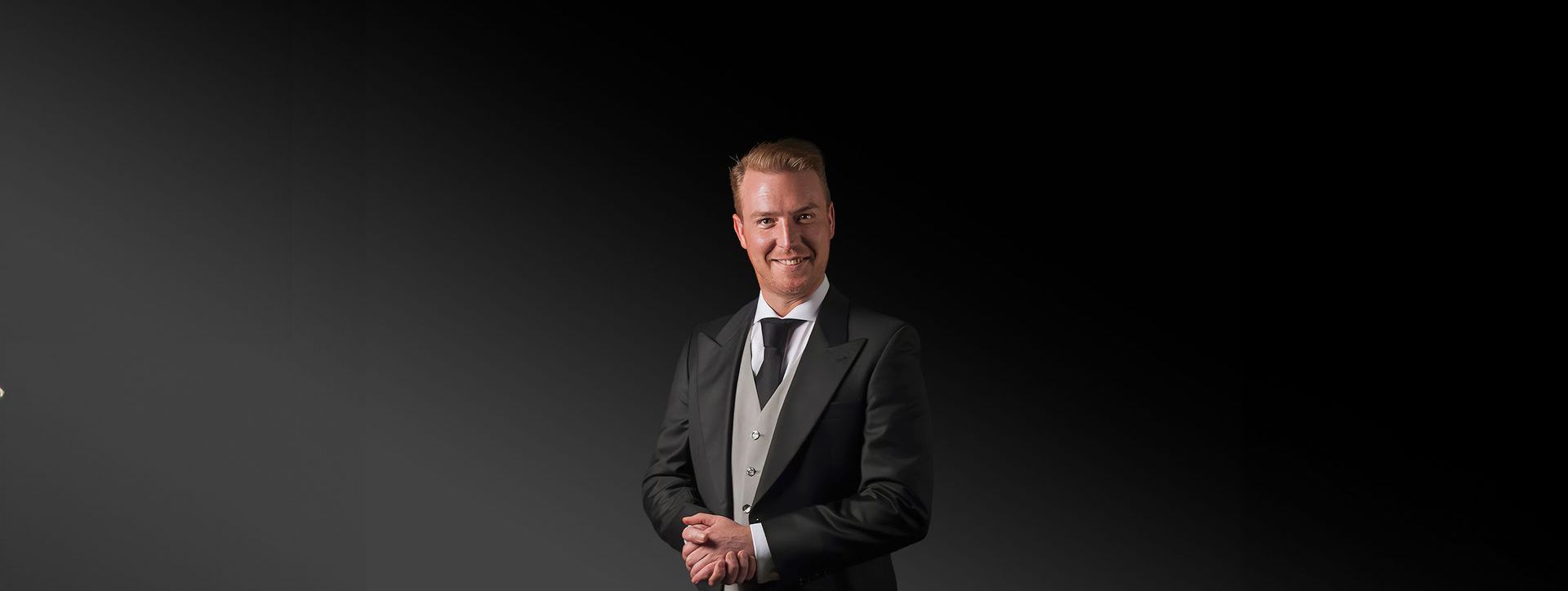 Een man in een smoking en stropdas staat voor een zwarte achtergrond.