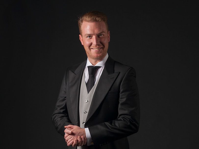 Een man in een smoking en stropdas staat met zijn handen gevouwen voor een zwarte achtergrond.