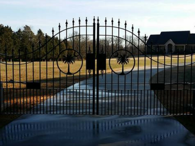 A wrought iron gate is open to a driveway with a house in the background