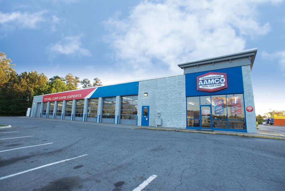 Exterior of an AAMCO Transmissions & Total Car Care shop with open bays, next to a parking lot and residential homes.