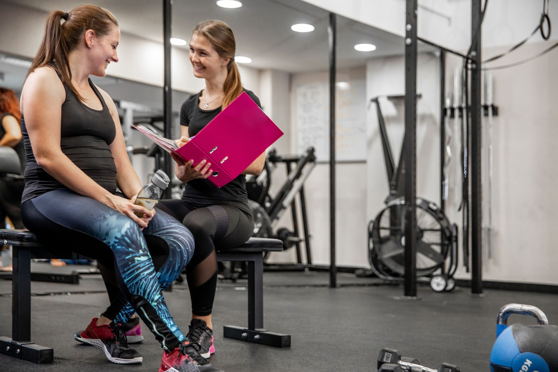 Two women are sitting on a bench in a gym talking to each other.