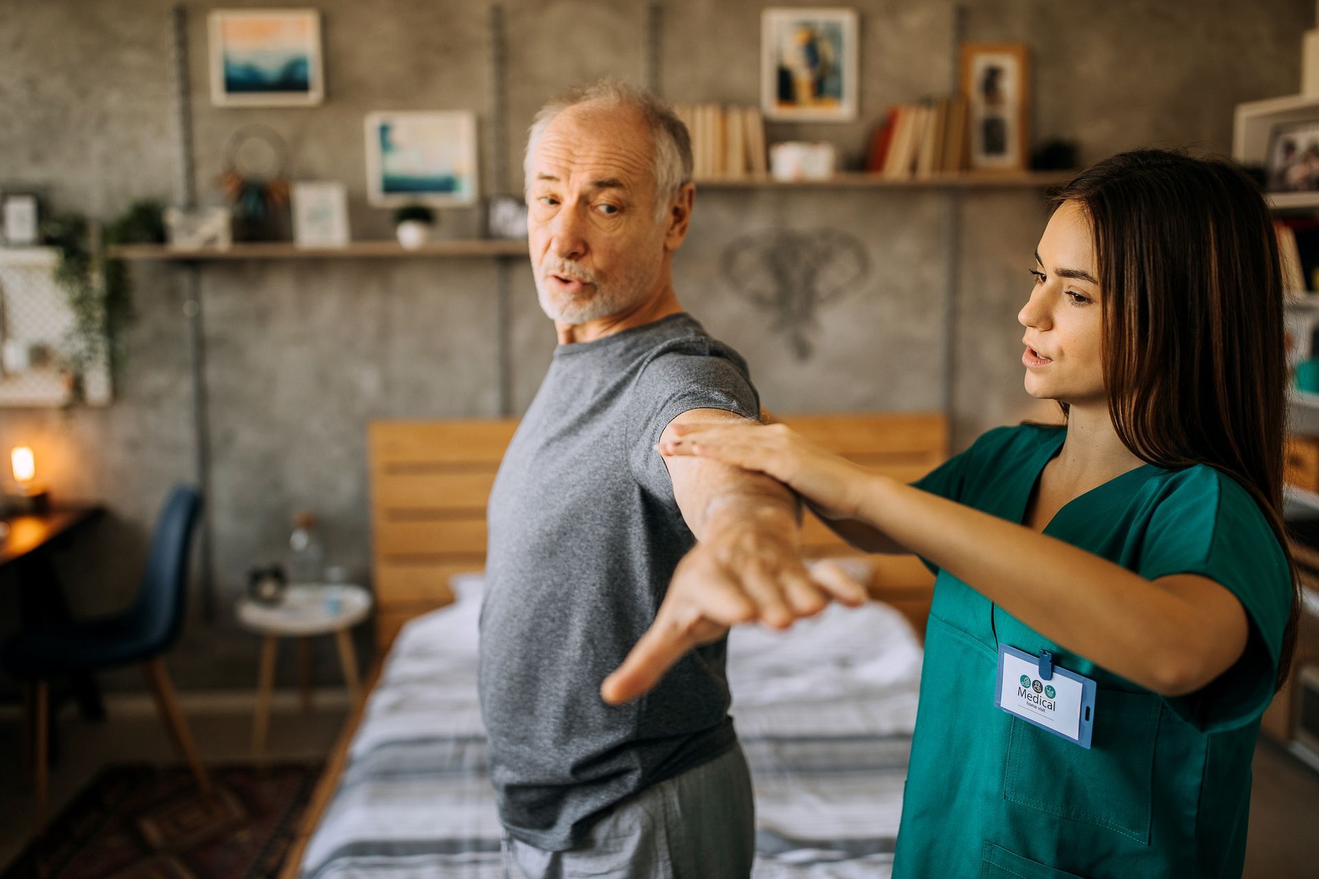 A nurse is helping an older man stretch his arms in a bedroom.