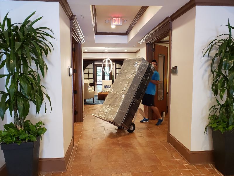A man is pushing a mattress on a dolly down a hallway.
