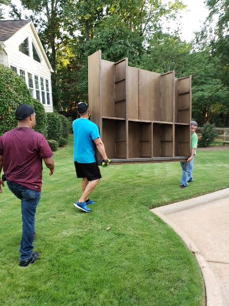 Two men are carrying a large wooden shelf in a yard.