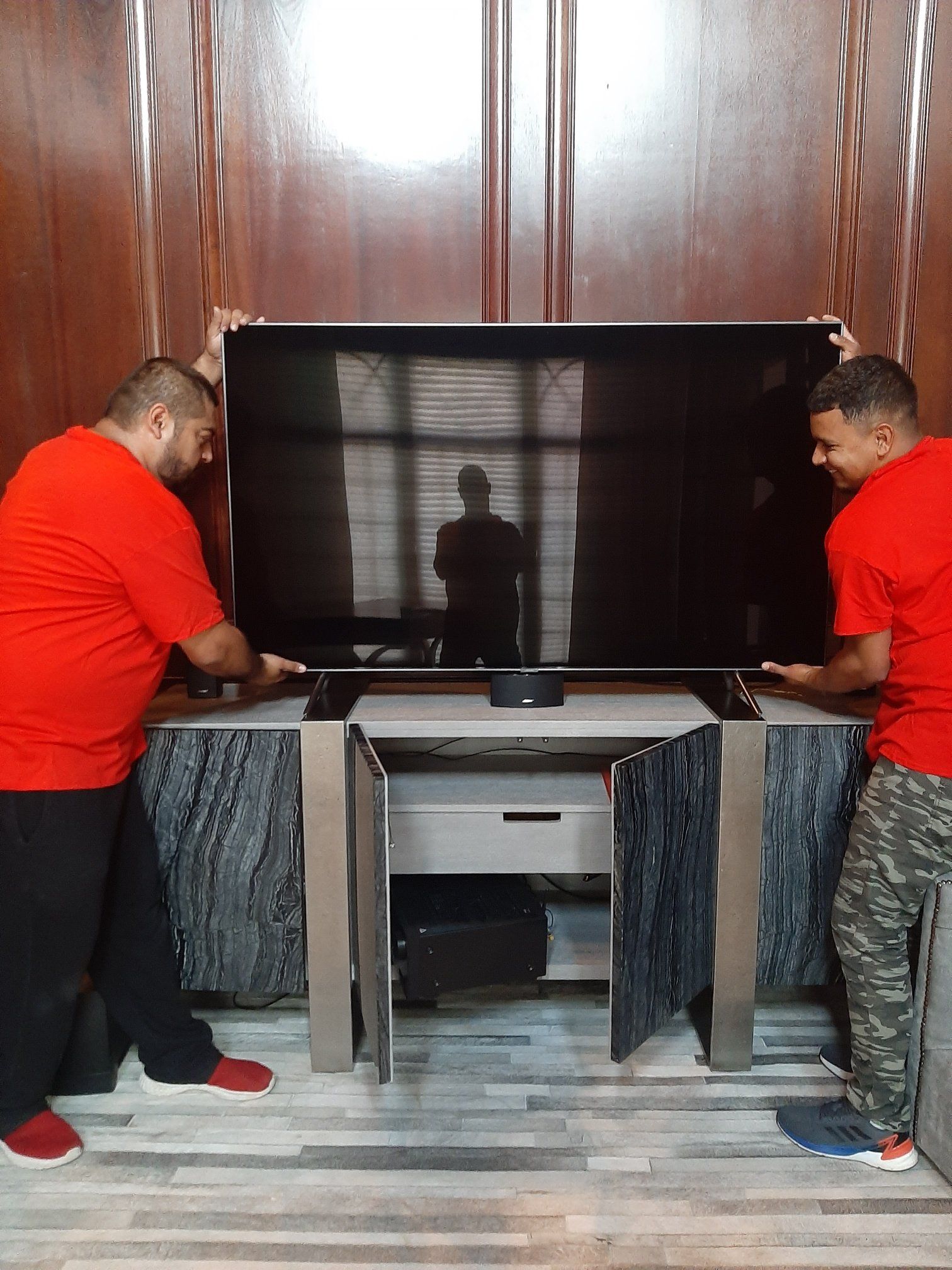 Two men in red shirts are holding a large flat screen tv.