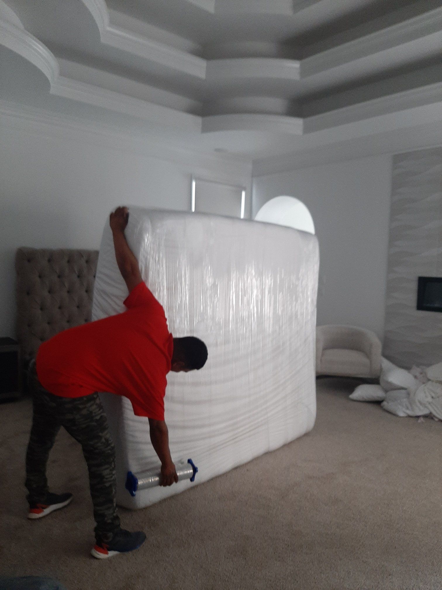 A man in a red shirt is wrapping a mattress in plastic wrap.