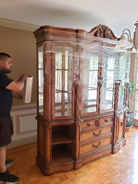 A man is wrapping a wooden cabinet with plastic wrap in a living room.