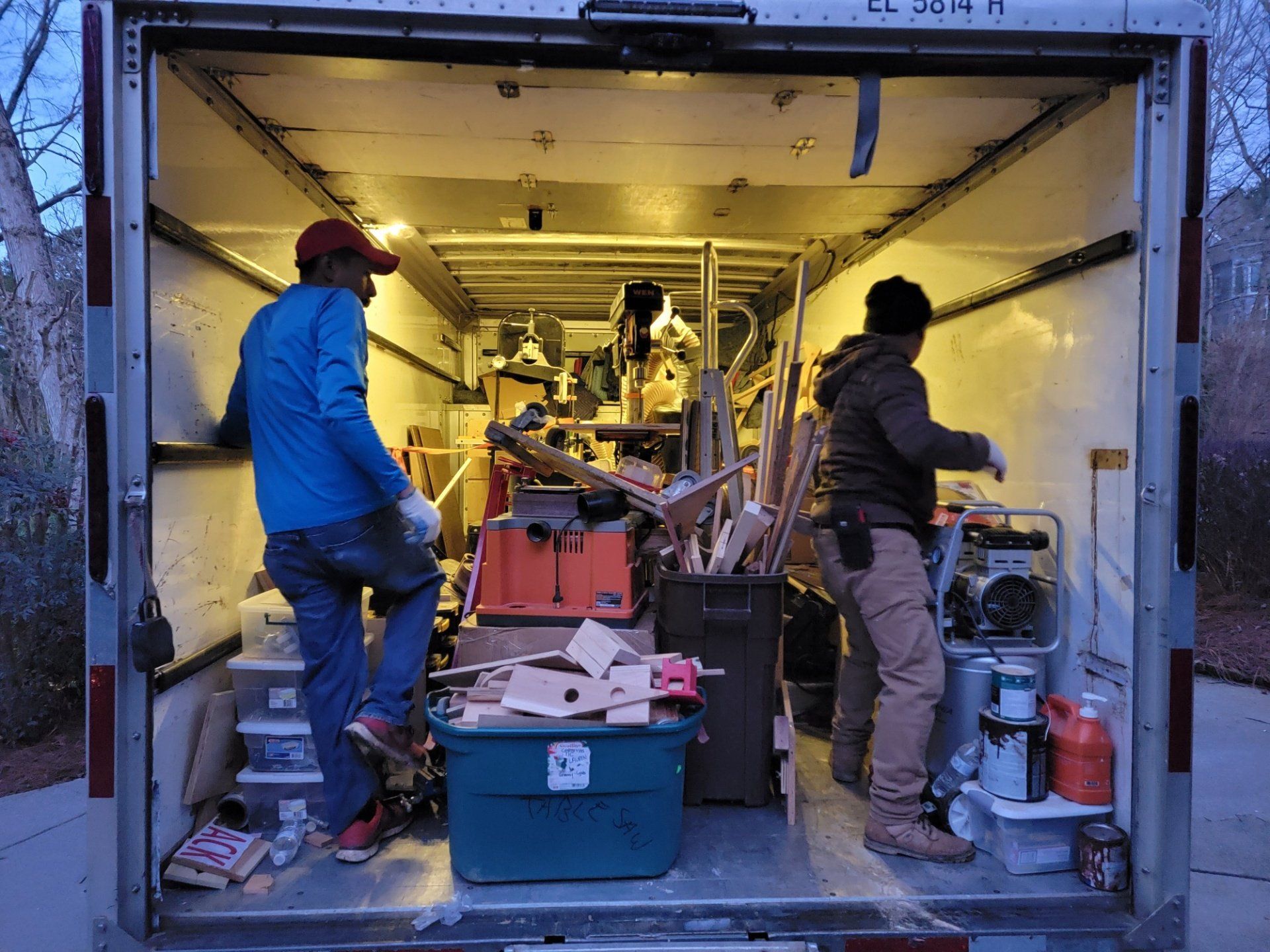 Two men are standing in the back of a trailer filled with junk.
