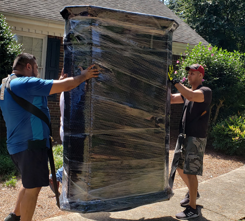 Two men are carrying a large refrigerator wrapped in plastic.