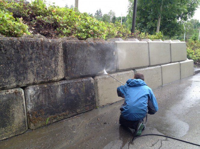 A man in a blue jacket is kneeling down and cleaning a stone wall with a high pressure washer.