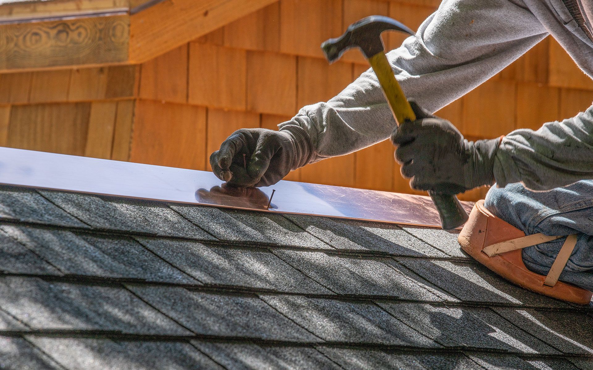 Roofer hammers copper flashing onto a shingled roof, wearing gloves.