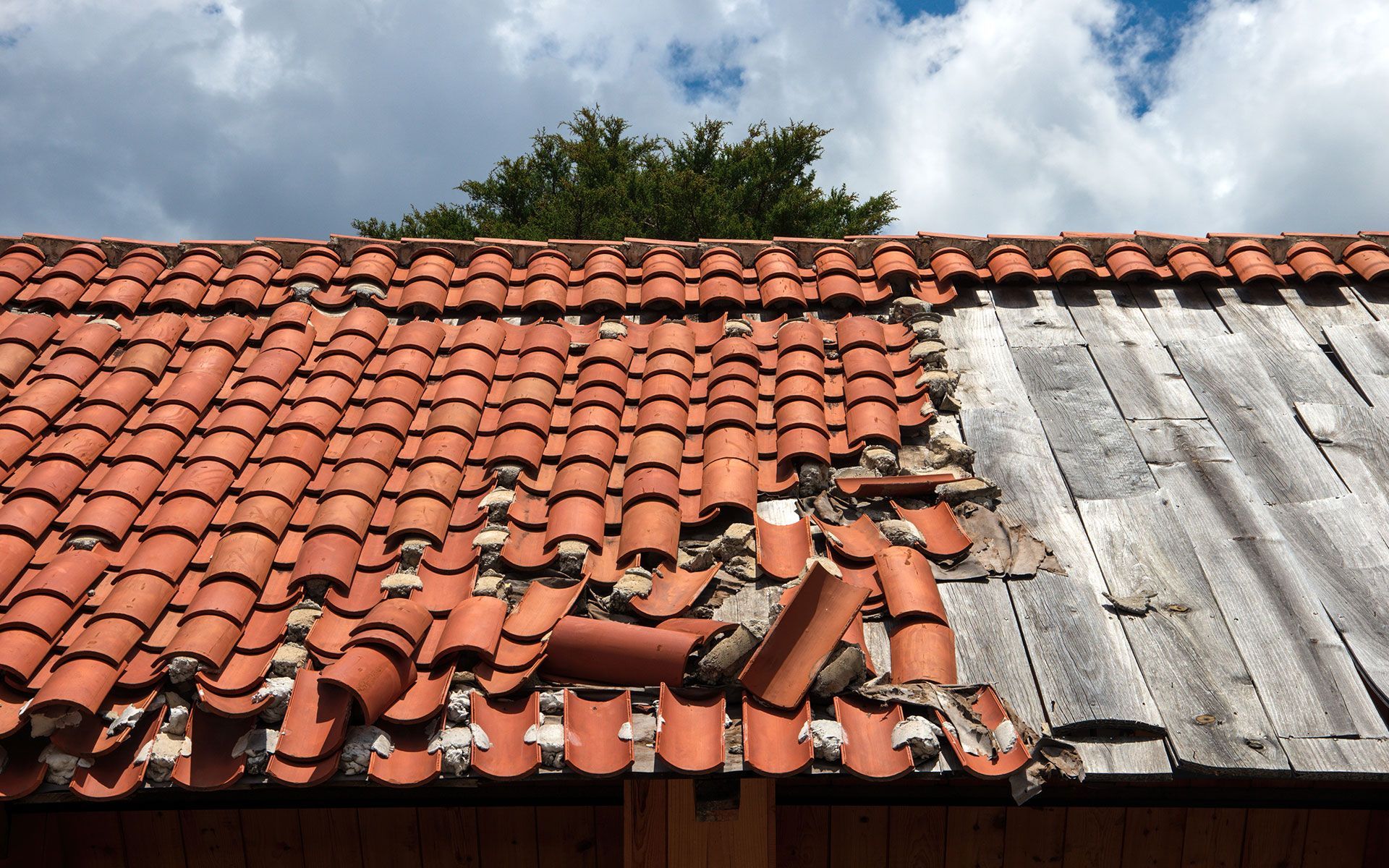 Damaged red tile roof with missing tiles, revealing weathered wood, against cloudy sky.