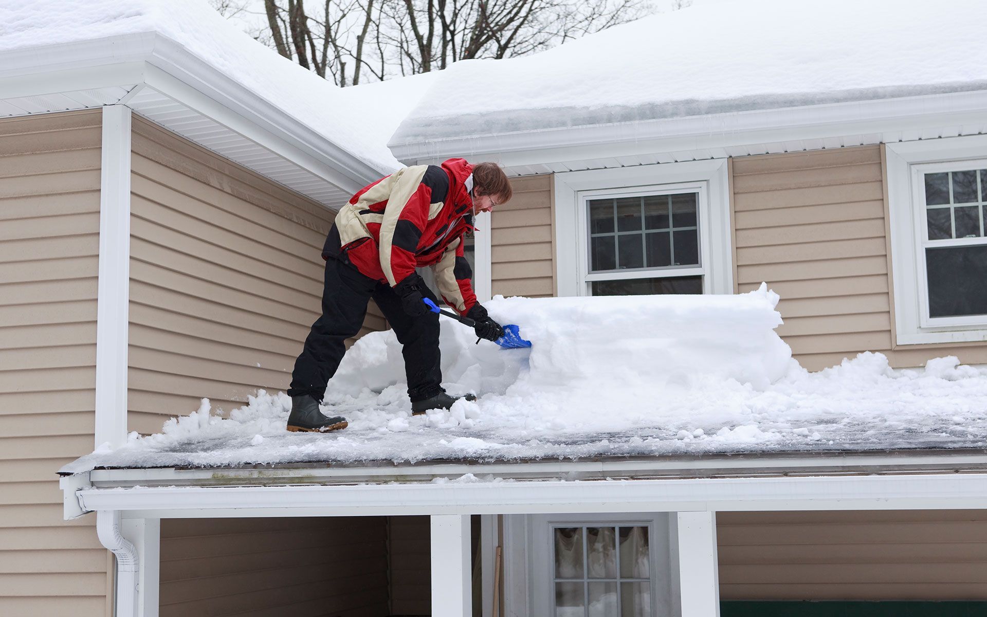 Man shoveling snow from a roof wearing a red jacket and black pants.