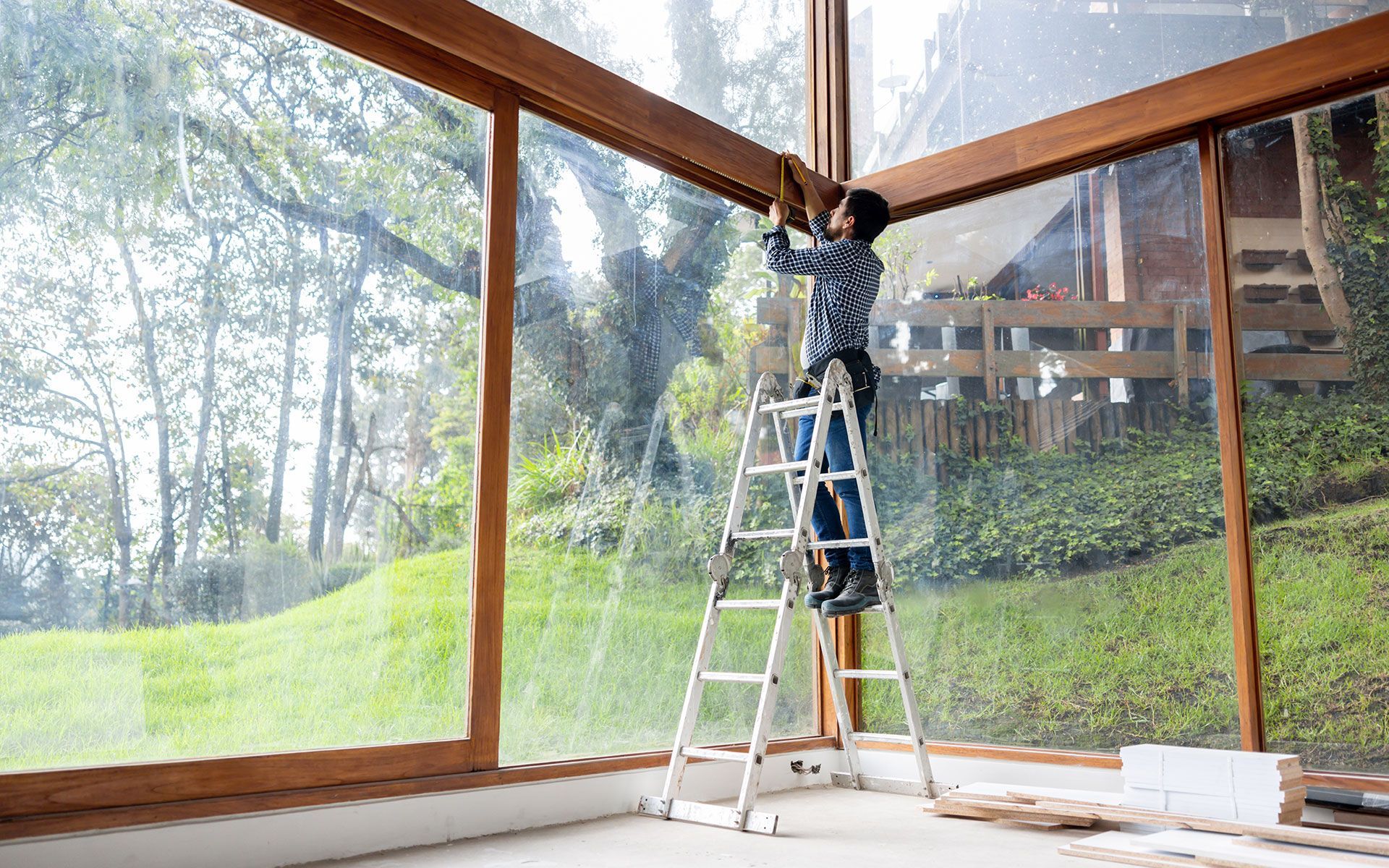 Person on a ladder installing trim around large windows in a room with a view of greenery.
