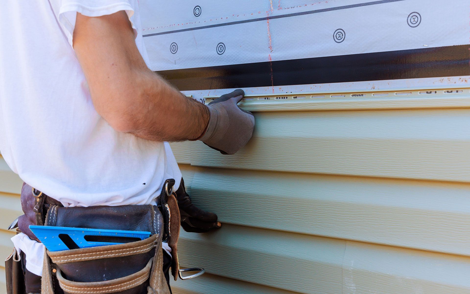A worker installing beige vinyl siding on a house, wearing gloves and a tool belt.