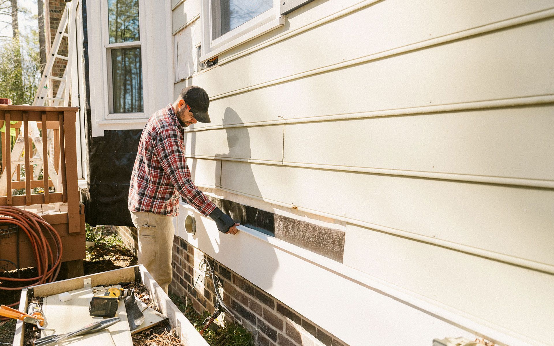 Man in plaid shirt repairs siding on a house, holding a tool near an opening.