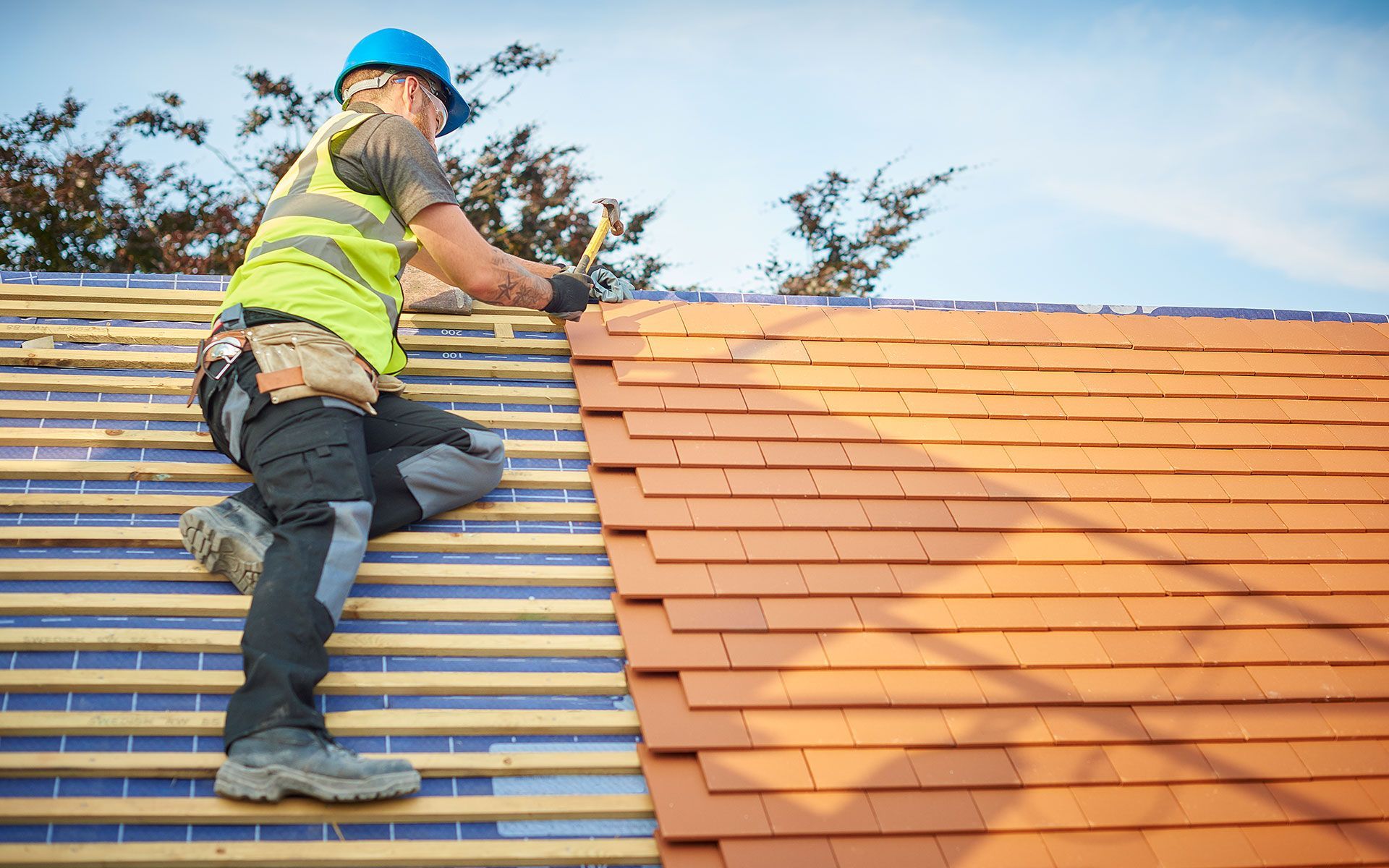 Roofer in blue hard hat installs orange roof tiles on a wooden frame under a blue sky.