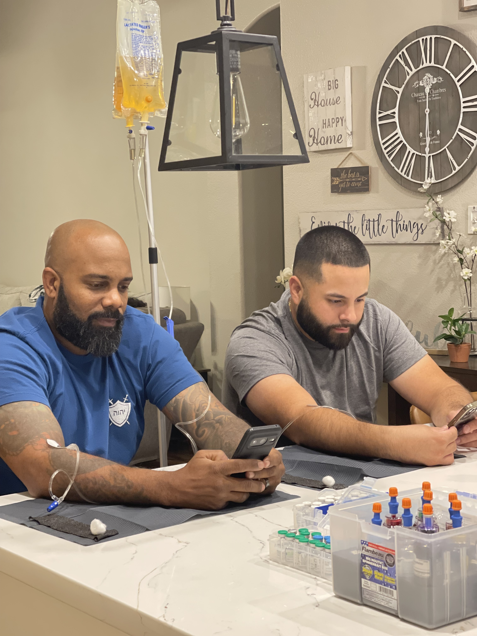 Two men are sitting at a table looking at their phones doing iv therapy
