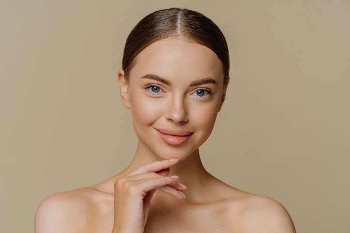 Headshot of beautiful brunette woman touches jawline looks directly at camera stands shirtless has natural beauty isolated over brown background. People wellness cosmetology skin care concept