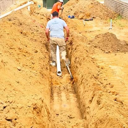 Two men working in a trench, installing white pipe. Sandy, outdoor construction site.