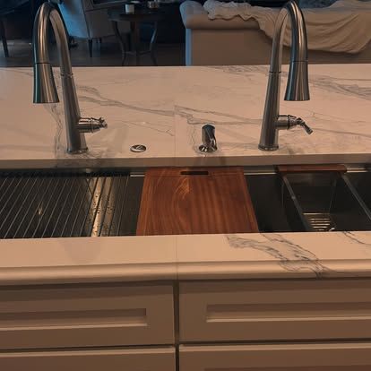 Kitchen sink with two faucets and a wooden cutting board. White cabinets and marble countertop.