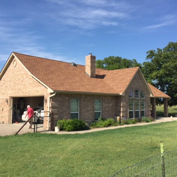 Brick house with brown roof, open garage, and a person carrying something outside on a sunny day.
