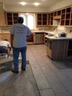 Two construction workers tiling a kitchen floor with exposed cabinets.