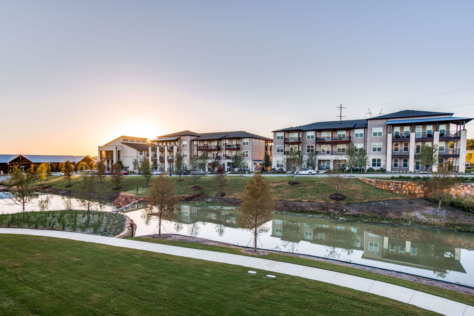 Apartment complex by a pond at sunset, with a walking path and green grass.