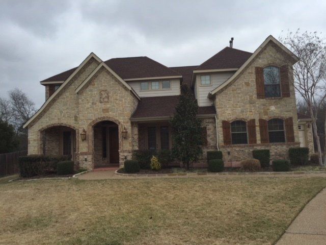 Two-story brick home with brown roof and shutters on a cloudy day; brown grass in front.