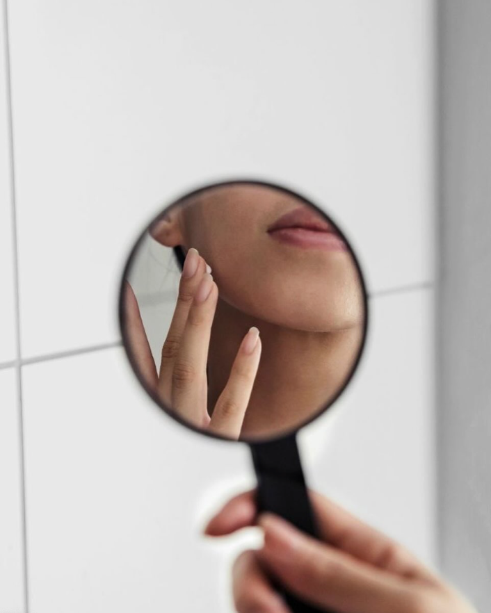 Person holding a round mirror, touching their face, close up of mouth and hand, white tiled wall.— Skin Haven Aesthetics Whitsundays in Airlie Beach, QLD
