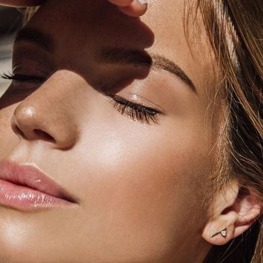 Close-up of a person with closed eyes, illuminated by sunlight. They're touching their forehead, with a small ear tattoo.— Skin Haven Aesthetics Whitsundays in Airlie Beach, QLD