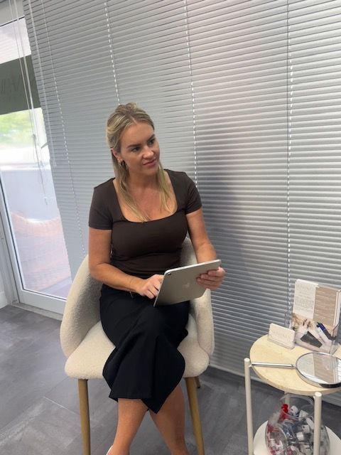 A blonde person wearing a brown top and black trousers sits in a chair, looking right while using a tablet — Skin Haven Aesthetics Whitsundays in Airlie Beach, QLD