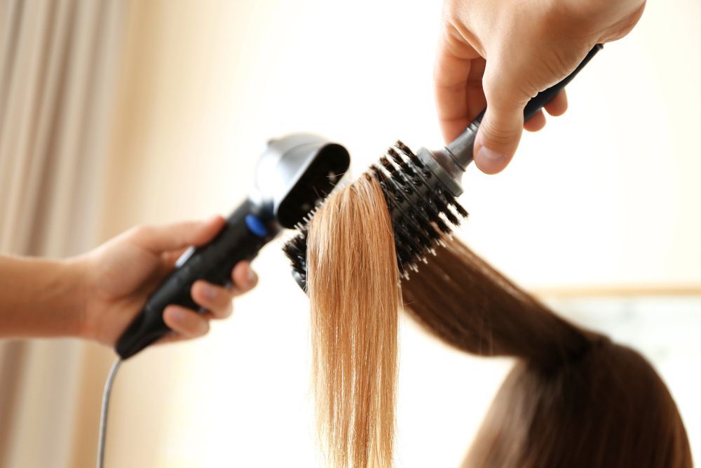 Una estilista utiliza un cepillo redondo y un secador de pelo para peinar el cabello castaño claro de una clienta.