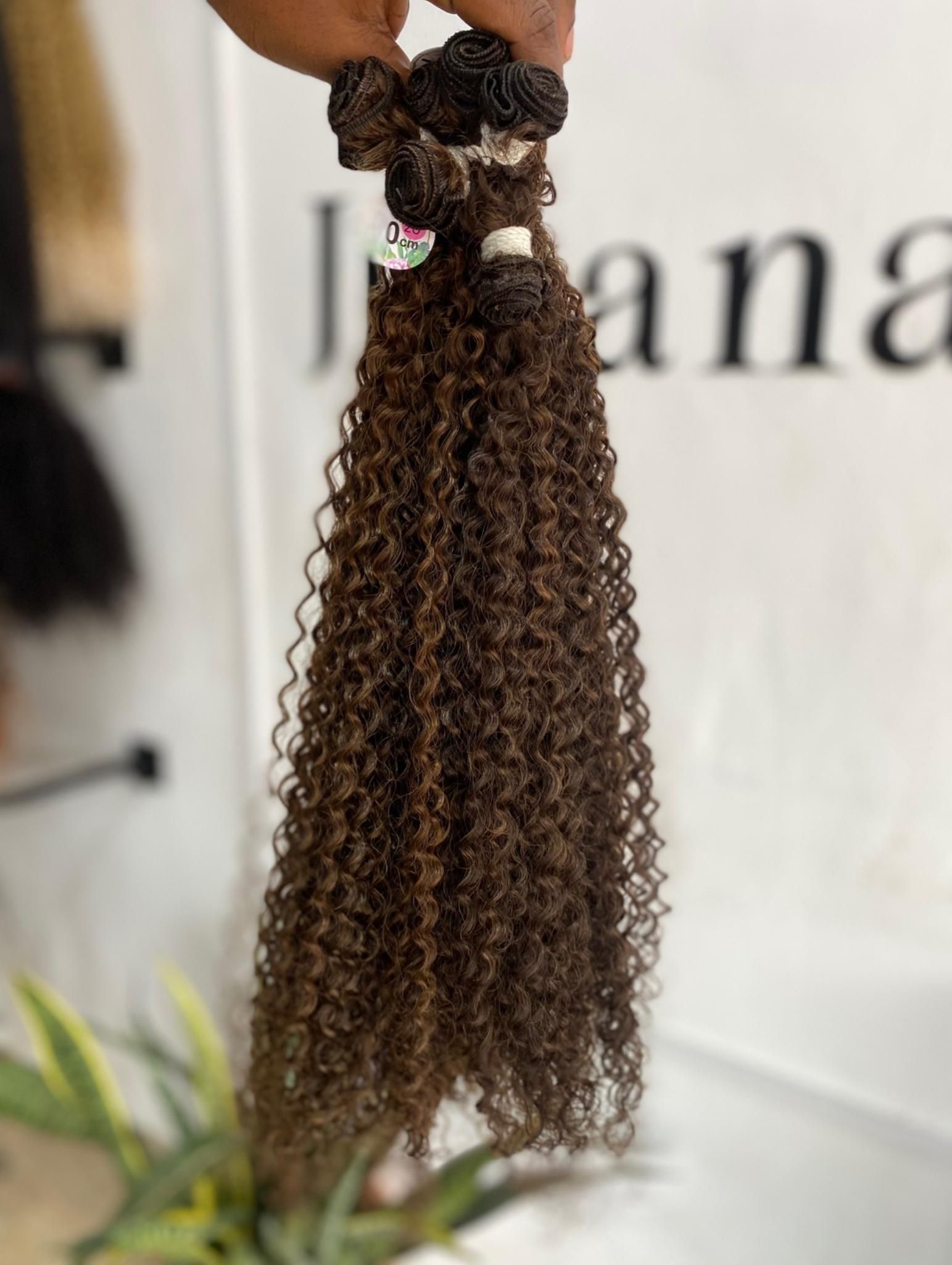 Una mano sostiene un mechón de extensiones de cabello rizado de color castaño oscuro sobre un fondo borroso con texto.