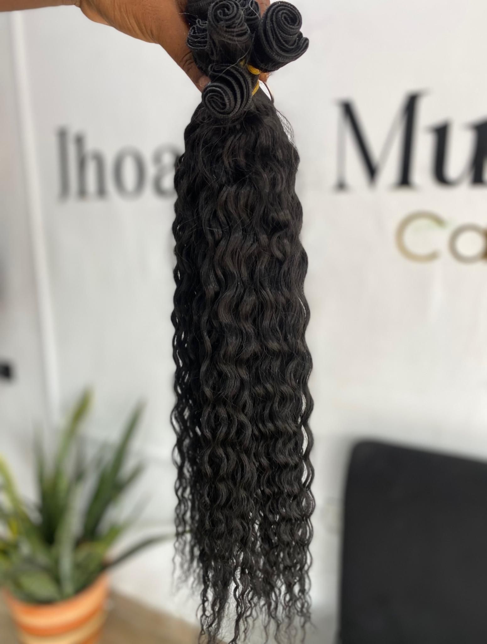 Una mano sostiene un manojo de extensiones de cabello oscuro y rizado frente a una pared blanca con un letrero parcial.