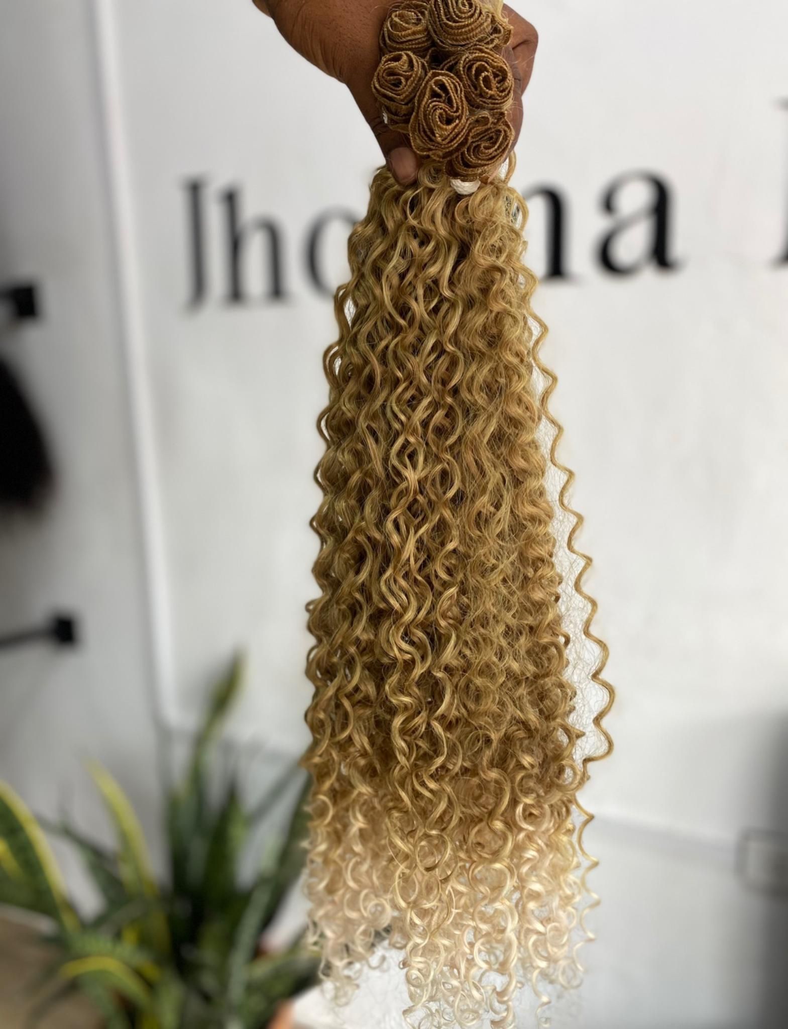 Una mano sostiene un mechón de extensiones de cabello rizado color rubio miel con un degradado rubio más claro en las puntas.