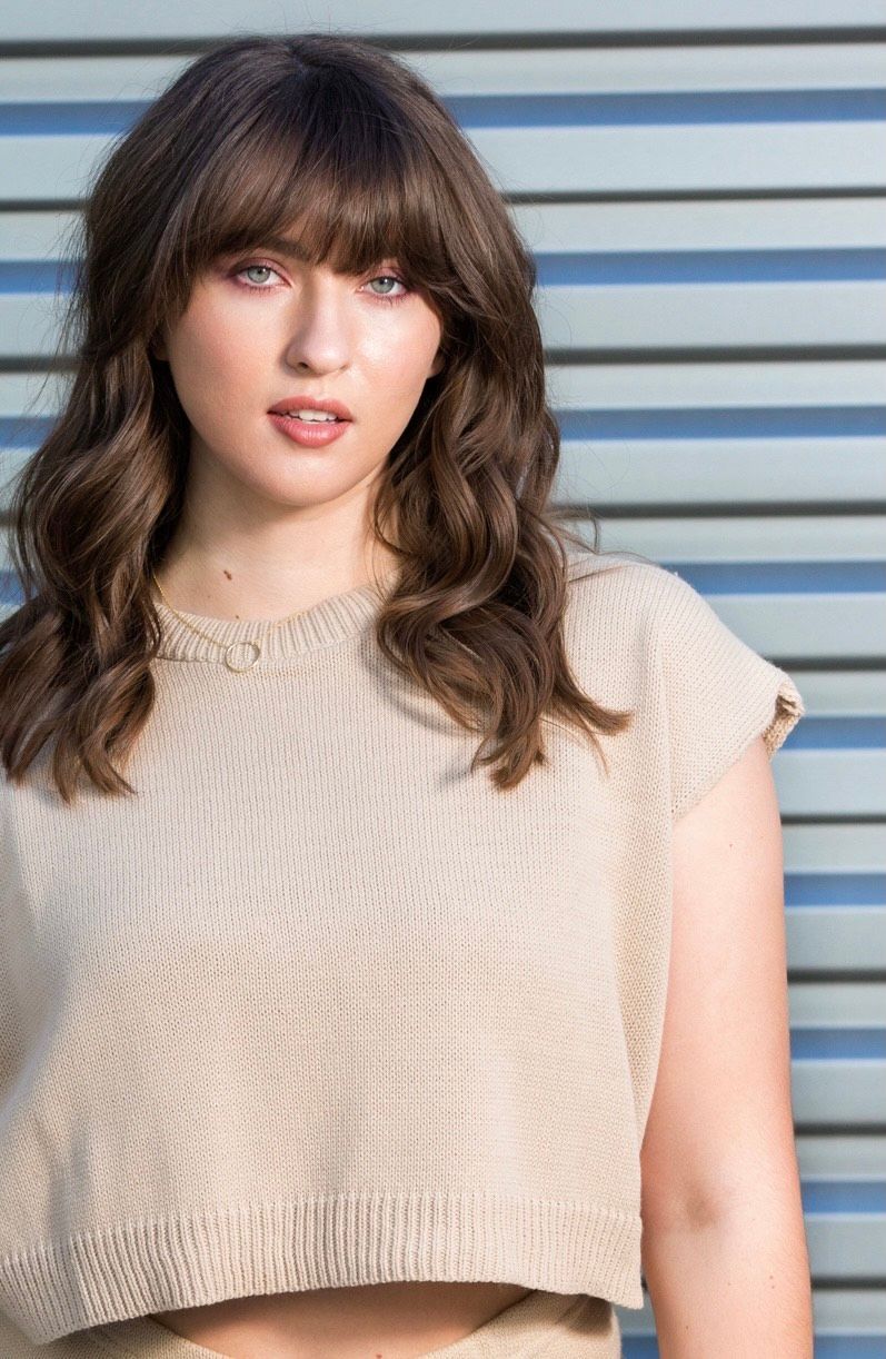 A woman wearing a crop top and a sweater is standing in front of a wall.