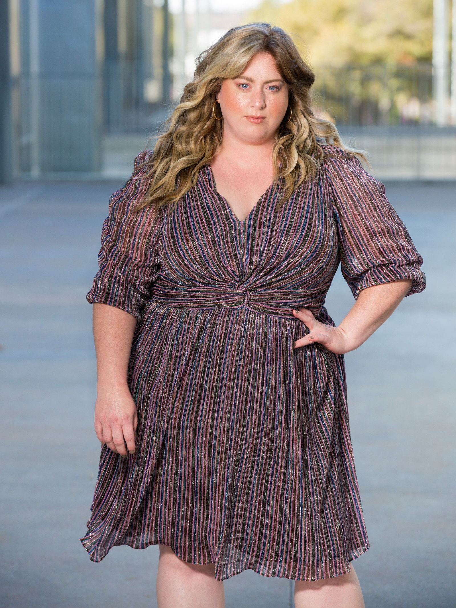 A woman in a dress is standing on a sidewalk with her hand on her hip.