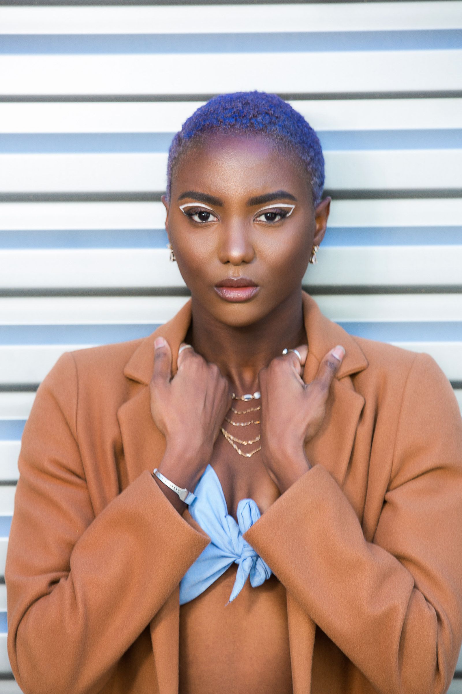 A woman with purple hair and a brown coat is standing in front of a wall.
