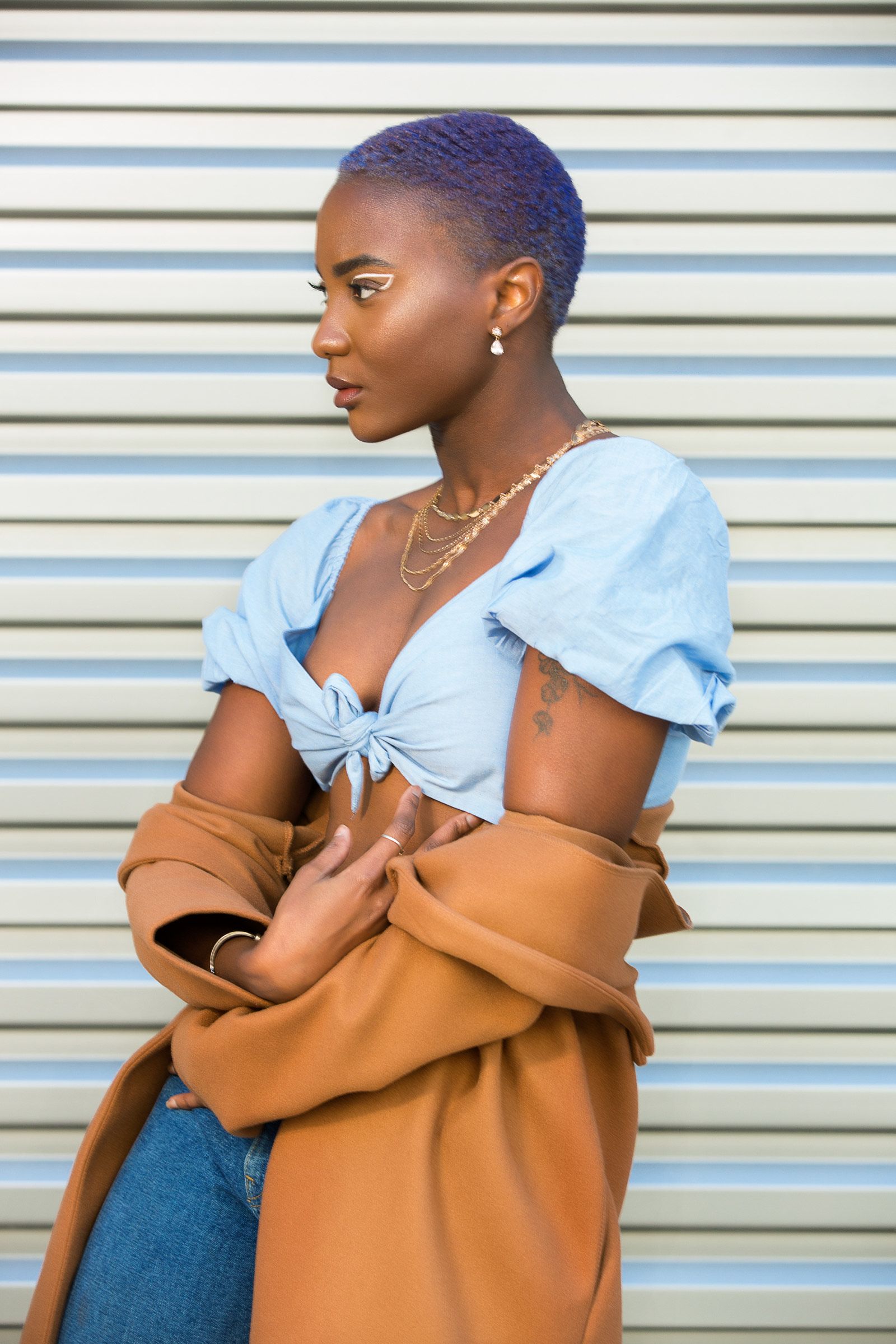 A woman with purple hair is wearing a blue top and a brown coat.
