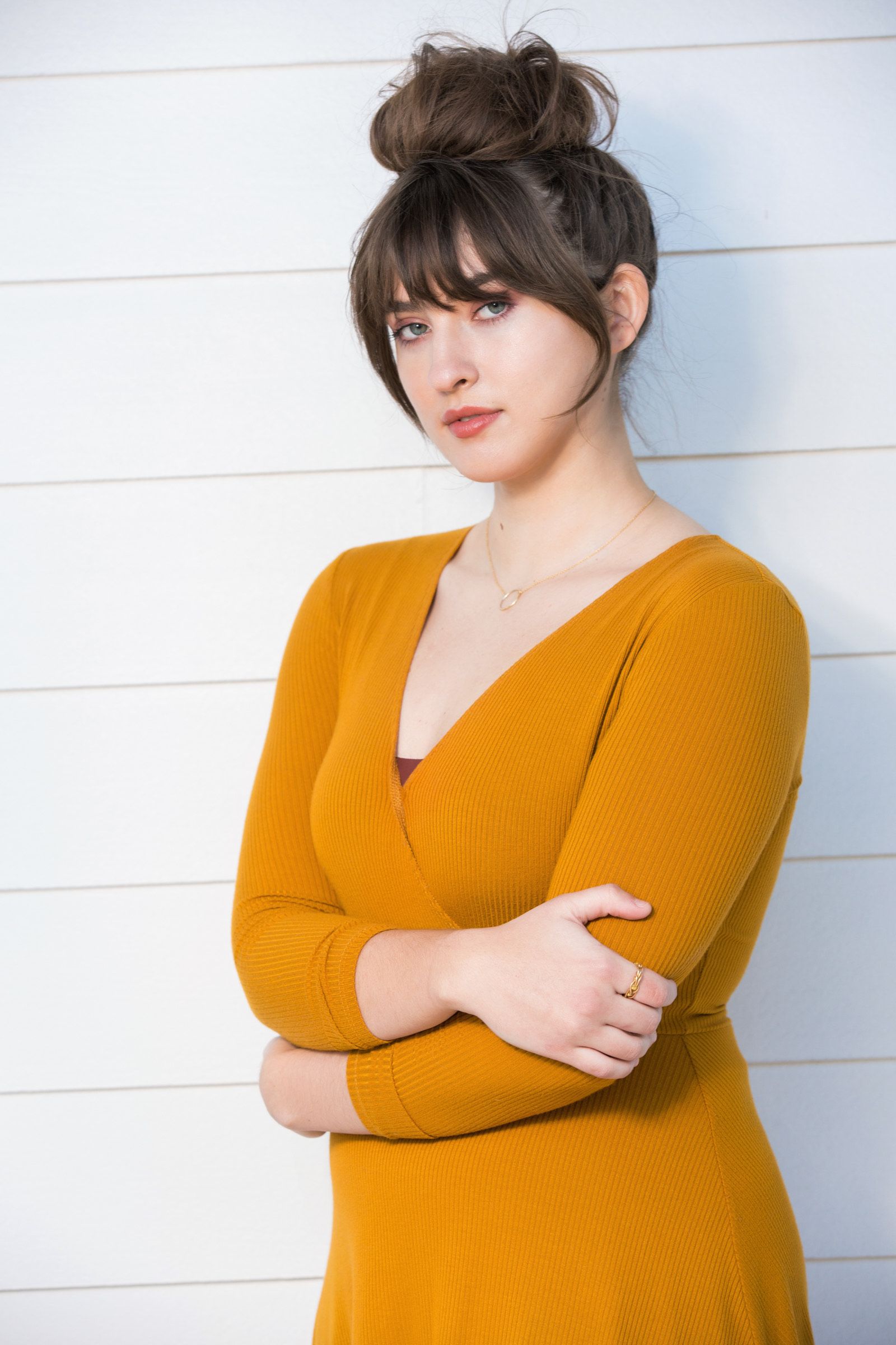 A woman in a yellow dress is standing in front of a white wall with her arms crossed.