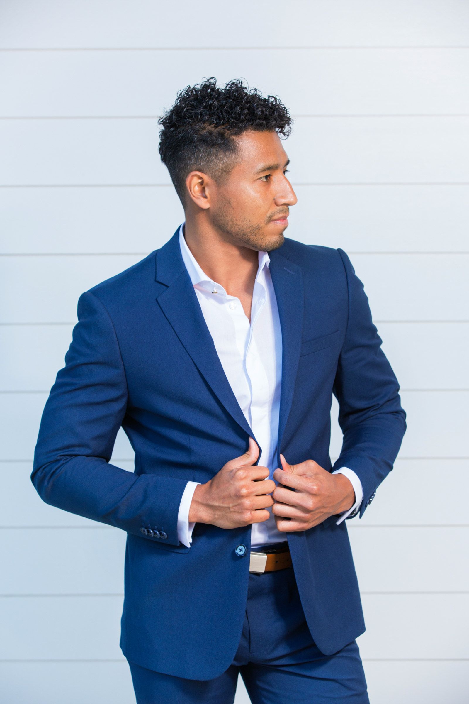 A man in a blue suit and white shirt is standing in front of a white wall.